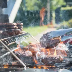 Des hamburgers grillent sur un barbecue à gaz. La fumée s'élève, les flammes dansent. Un hamburger est retourné avec une pince.