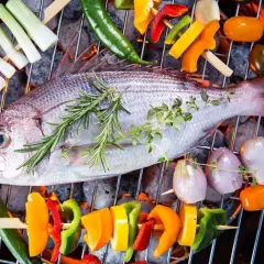 Poisson entier sur un barbecue électrique, entouré de brochettes de légumes colorés et d'herbes aromatiques.