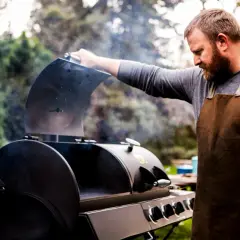 Un homme barbu en tablier ouvre un fumoir d'où s'échappe de la fumée. Il est prêt à apprendre comment allumer un fumoir pour un repas savoureux.
