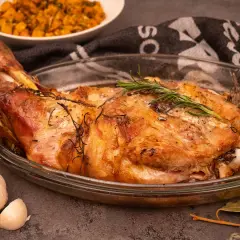 Une succulente épaule d'agneau rôtie, garnie de romarin frais, dans un plat en verre. A côté, de l'ail et une purée de légumes.
