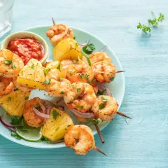 Brochettes de scampis grillés avec des morceaux d'ananas, de la ciboulette et une sauce rouge épicée sur une assiette bleue.