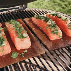 Quatre pavés de saumon sur planche de cèdre, garnis d'herbes fraîches et d'ail, prêts à être grillés.