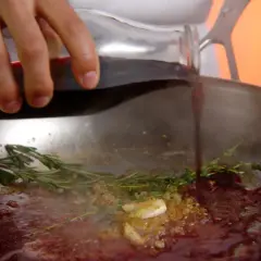 Une main verse du jus de viande dans une poêle avec de l'ail et des herbes.