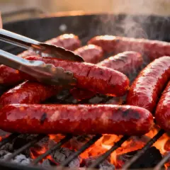 Des merguez juteuses grésillent sur un barbecue, dégageant une fumée alléchante. Un geste expert les retourne avec des pinces pour une cuisson parfaite.
