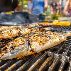 Trois poissons entiers, marqués par le barbecue, grillent sur une grille. Des éclats de citron sont visibles en arrière-plan.