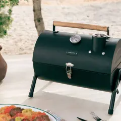 Barbecue noir avec couvercle, thermomètre et poignée en bois, prêt pour le temps de cuisson. Une carafe en terre cuite et une assiette de tomates colorées sont à proximité.