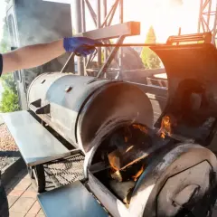 Un homme ajuste le bois pour fumoir dans un grand barbecue. Des flammes dansent dans le foyer, promettant une cuisson savoureuse.