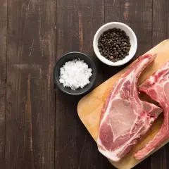Deux belles côtes de porc crues sur une planche à découper en bois, accompagnées de sel et de grains de poivre noir.