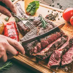 Une main tranche une côte de bœuf juteuse, résultat d'une cuisson au barbecue charbon parfaite. Accompagnée de pommes de terre et poivrons grillés.