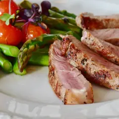Tranches de secreto de porc grillé, accompagnées d'asperges vertes et de tomates cerises sur une assiette blanche.