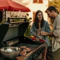 Un couple prépare une soiree barbecue party. Des steaks cuisent sur le grill pendant qu'ils coupent des légumes.