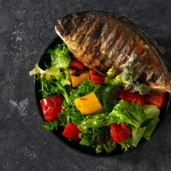 Dorade au barbecue, entière et grillée, servie sur un lit de salade verte, poivrons rouges et jaunes, et brocolis.