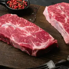 Deux beaux steaks de l angus beef, marbrés de gras, prêts à être cuisinés sur une planche en bois. Poivre rose en accompagnement.