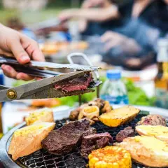 Une main coupe un steak juteux sur un gril lors d'une fête barbecue. Des pains grillés, des pommes de terre et du maïs complètent le festin.