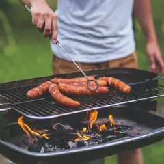 Un homme retourne des saucisses sur un barbecue bois ou charbon. Les flammes dansent sous la grille, promettant un repas savoureux.