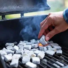 Une main allume des briquettes de charbon dans un barbecue, de la fumée s'échappe. On se demande combien de temps pour allumer un barbecue.
