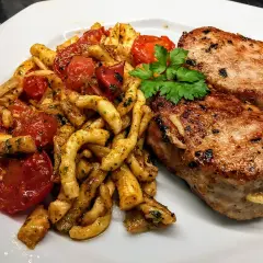 Deux côtelettes de veau grillées, servies avec des pâtes torsadées et des tomates cerises poêlées, garnies de persil frais. Un plat savoureux et coloré.