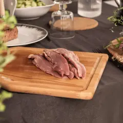 Aiguillettes de canard crues sur une planche à découper en bois, prêtes à être cuisinées. Des herbes fraîches et du pain complètent la scène.