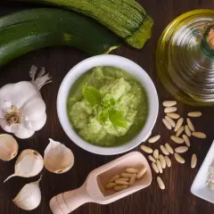 Un bol de pesto de courgettes frais, garni de basilic, entouré d'ingrédients : courgettes, ail, pignons de pin, parmesan râpé et huile d'olive.