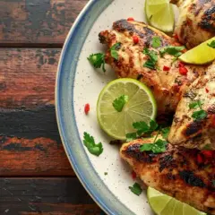 Poulet grillé, garni de coriandre fraîche, de piment rouge et de quartiers de citron vert, évoquant une savoureuse marinade mexicaine.