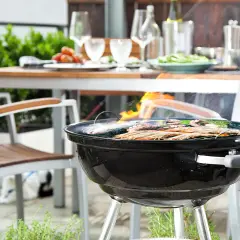 Un barbecue au charbon est allumé, avec des crevettes et du poisson grillant sur la grille. Une table est dressée à l'arrière, prête pour un repas.
