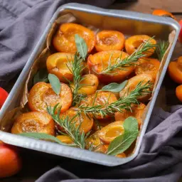 Abricots rôtis dans un plat, parsemés de romarin et de sauge. Des fruits frais entiers et coupés entourent le plat.