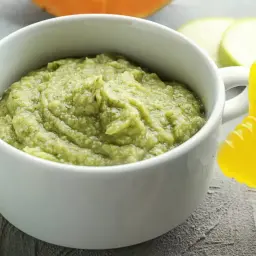 Un bol de purée de courgette onctueuse, à côté d'une autre purée orangée et d'une rondelle de courgette. Un jouet de dentition jaune est aussi visible.