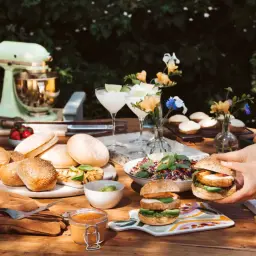 Préparation d'une recette pique nique : burgers, salade colorée, fruits et boissons rafraîchissantes sur une table en bois.