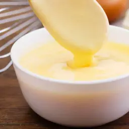 Une cuillère verse une sauce hollandaise onctueuse dans un bol blanc. Un fouet et des ingrédients de cuisine sont visibles en arrière-plan.