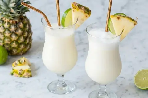 Deux verres de pina colada, décorés d'ananas et de citron vert, avec des pailles dorées. Un ananas entier et des citrons verts sont disposés autour.