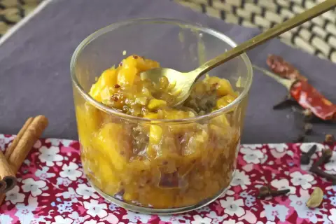 Un verre rempli de chutney de mangue, avec une cuillère dorée. Des épices comme la cannelle, le piment et le clou de girofle entourent le plat.