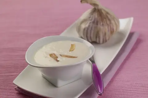 Un bol de crème d'ail onctueuse, garnie de fines lamelles d'ail croustillantes, à côté d'une tête d'ail fraîche.
