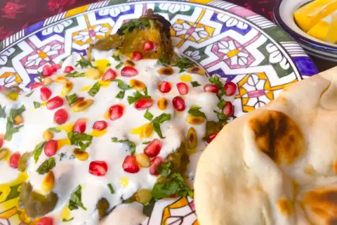 Un plat de mutabal crémeux, garni de grenades et de pignons, servi avec du pain pita sur une assiette décorée.