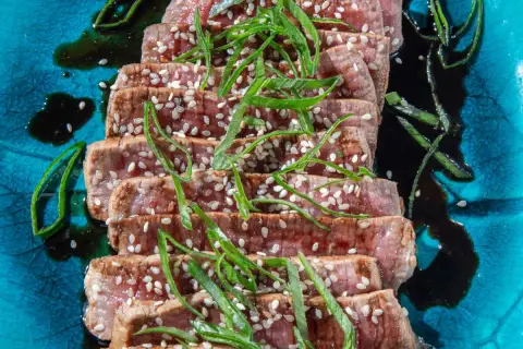 Tranches de tataki de boeuf, parsemées de graines de sésame et garnies de ciboule, sur une assiette bleue texturée.