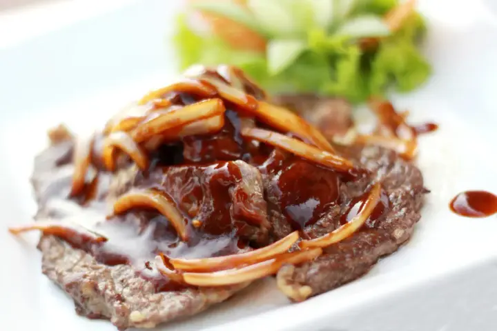 Un steak savoureux nappé d'une sauce brune et d'oignons caramélisés, servi avec une garniture de salade verte.