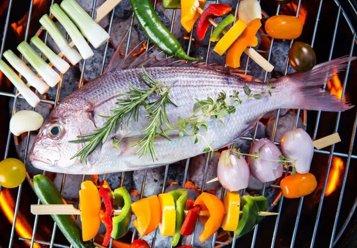 Poisson entier sur un barbecue électrique, entouré de brochettes de légumes colorés et d'herbes aromatiques.