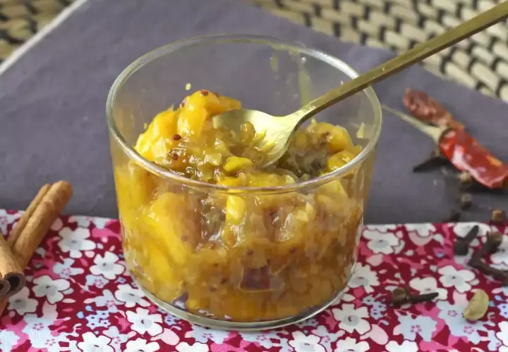 Un verre rempli de chutney de mangue, avec une cuillère dorée. Des épices comme la cannelle, le piment et le clou de girofle entourent le plat.