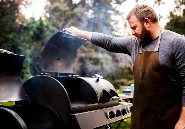 Un homme barbu en tablier ouvre un fumoir d'où s'échappe de la fumée. Il est prêt à apprendre comment allumer un fumoir pour un repas savoureux.