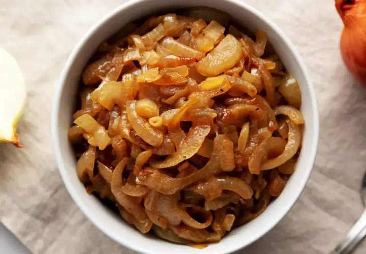 Un bol rempli d'oignons caramélisés dorés, parfaits pour une recette savoureuse. Un oignon cru à côté.
