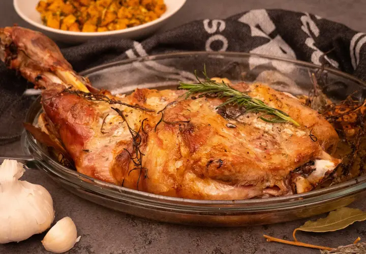 Une succulente épaule d'agneau rôtie, garnie de romarin frais, dans un plat en verre. A côté, de l'ail et une purée de légumes.
