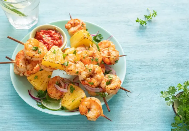 Brochettes de scampis grillés avec des morceaux d'ananas, de la ciboulette et une sauce rouge épicée sur une assiette bleue.