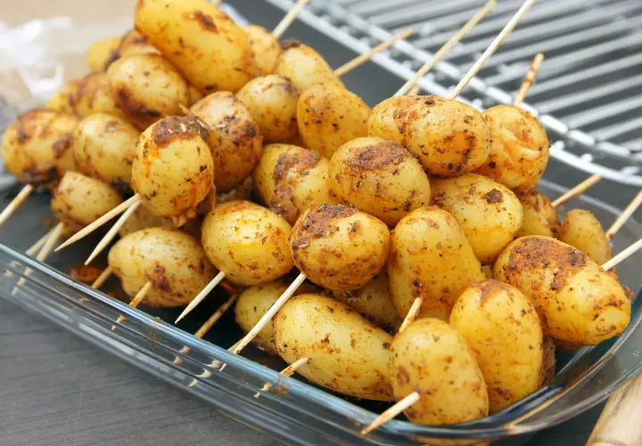 Des brochettes de pommes de terre assaisonnées, prêtes pour le barbecue, dans un plat en verre.
