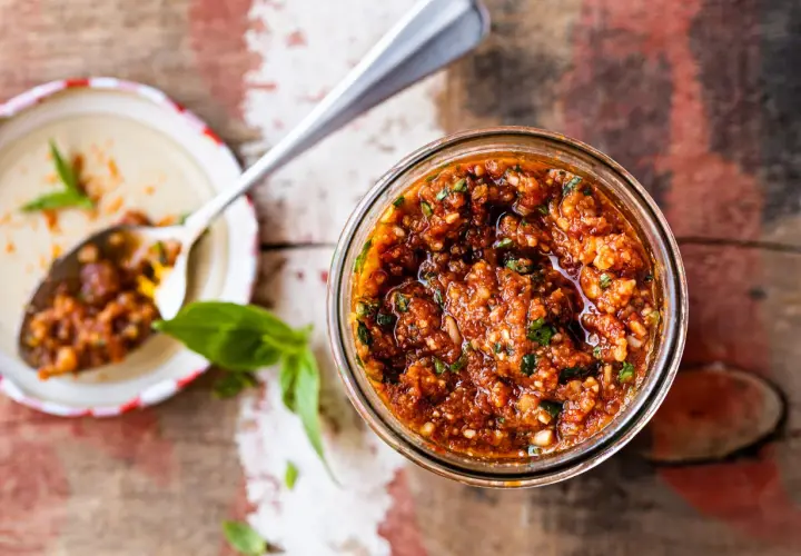 Un pot de pesto rosso, aux tomates séchées et basilic, prêt à être dégusté. Une cuillère en argent en est remplie.