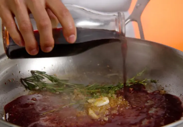 Une main verse du jus de viande dans une poêle avec de l'ail et des herbes.
