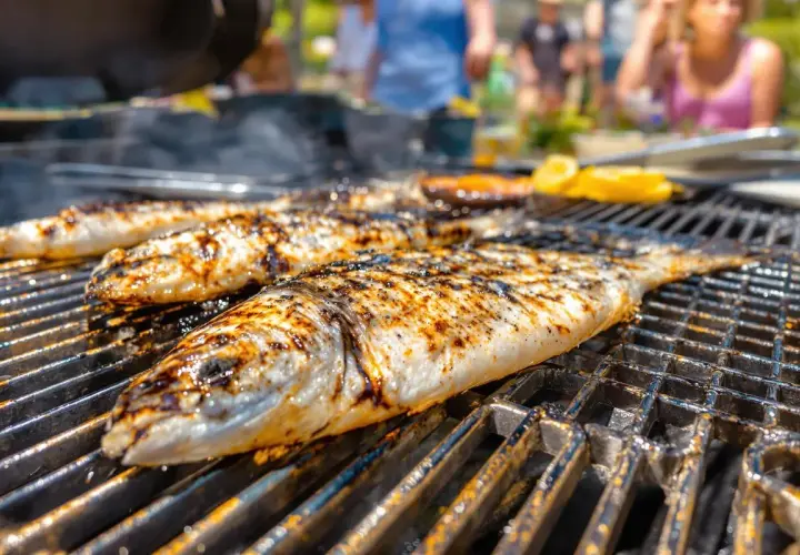Trois poissons entiers, marqués par le barbecue, grillent sur une grille. Des éclats de citron sont visibles en arrière-plan.