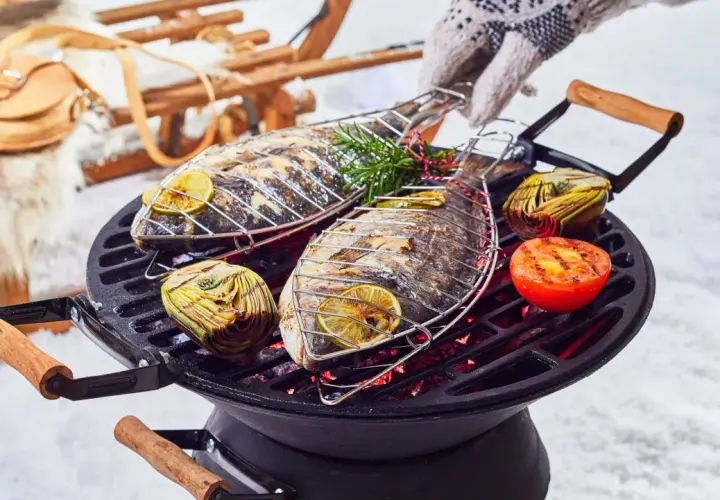 Deux poissons entiers grillent sur un barbecue, accompagnés d'artichauts et de tomates. Un gant tricoté ajuste la grille. Un vrai barbecue hivernal !