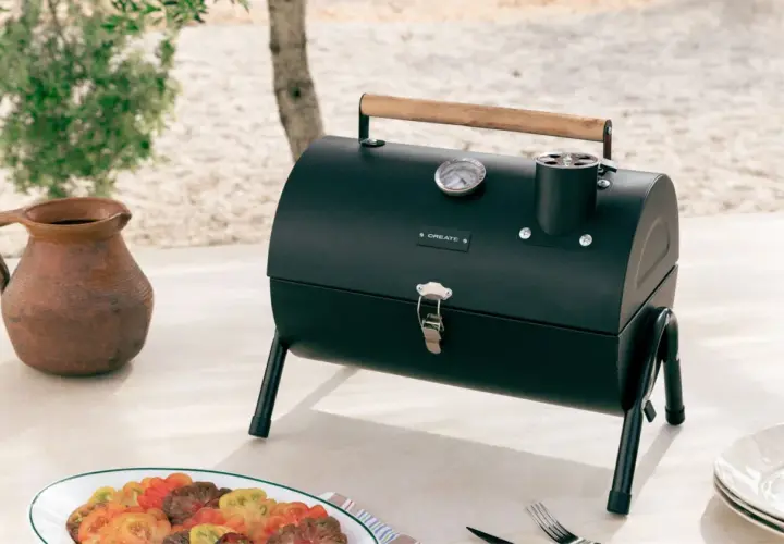 Barbecue noir avec couvercle, thermomètre et poignée en bois, prêt pour le temps de cuisson. Une carafe en terre cuite et une assiette de tomates colorées sont à proximité.