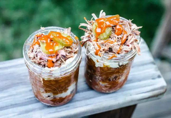Deux pots en verre remplis de pulled pork, haricots et sauce barbecue, parfaits pour un brunch barbecue en plein air.