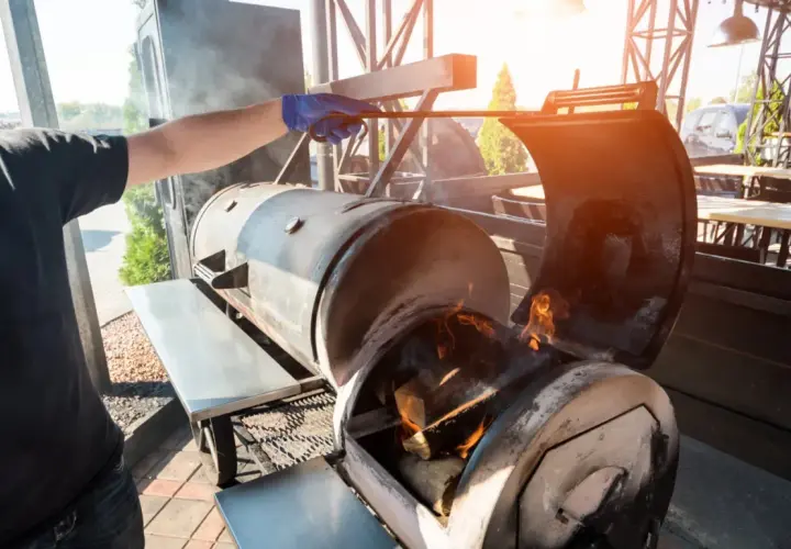 Un homme ajuste le bois pour fumoir dans un grand barbecue. Des flammes dansent dans le foyer, promettant une cuisson savoureuse.