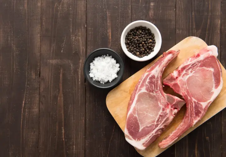 Deux belles côtes de porc crues sur une planche à découper en bois, accompagnées de sel et de grains de poivre noir.